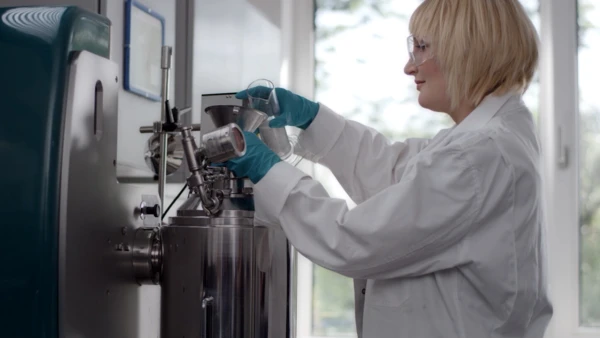 Eine Technikerin in einem Labor mit Handschuhen fügt Materialien in eine Mischmaschine ein, fokussiert auf präzise Verarbeitungstechniken.